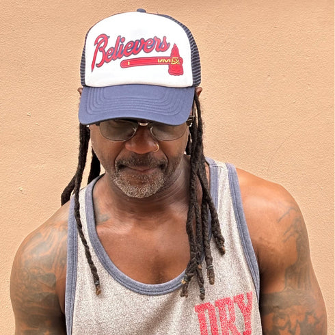 Man wearing a cap with 'Believers' text against a beige background
