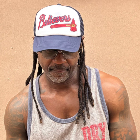 Man wearing a cap with 'Believers' text against a beige background