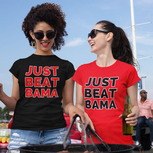 Two women outside tailgating wearing 'Just Beat Bama' t-shirts, one shirt is red, and the other shirt is black.