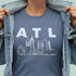 Person wearing a blue t-shirt with 'ATL' and city skyline graphic, holding denim jacket open.